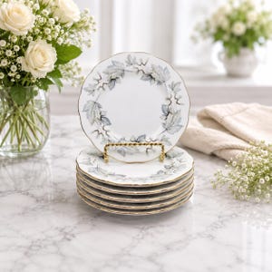Puede incluir: Seis platos de porcelana blanca con borde dorado y diseño de hojas en tonos grises y azules. Los platos se exhiben en un soporte dorado. Un ramo de rosas blancas en el fondo. Cada plato mide aproximadamente 20 cm de diámetro.