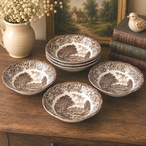 May include: A set of four ceramic bowls featuring a brown and white transferware pattern depicting a rural landscape with trees and rolling hills. Each bowl has a scalloped rim and a detailed floral border, showcasing a consistent pastoral scene.