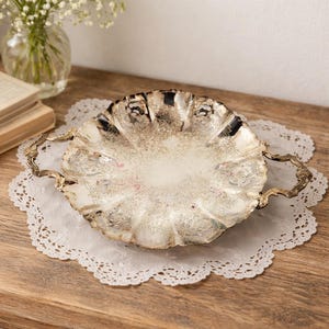 May include: Silver-plated serving tray with a scalloped rim and decorative handles. The tray has detailed embossed patterns and rests on a white lace doily. A vintage piece, ideal for serving or display purposes.