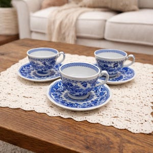 May include: Three blue and white porcelain teacups and saucers with a floral pattern are displayed on a wooden table. The teacups have a classic shape with a handle, and the saucers are round. The set is placed on a cream-coloured lace doily.