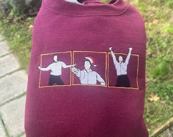 Op de afbeelding: Bordeauxkleurige trui met een geborduurd beeld van drie figuren in een kerstscène. De figuren dragen witte shirts en zwarte broeken, en één figuur draagt een kerstmanmuts. De figuren zijn omlijst met goud.