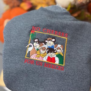 May include: A dark gray sweatshirt with an embroidered design. The design features a group of cartoon mice inside a gold rectangle, with the text "NO CHEESES FOR US MEECE" in red.