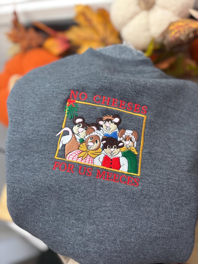 May include: A dark gray sweatshirt with an embroidered design. The design features a group of cartoon mice inside a gold rectangle, with the text "NO CHEESES FOR US MEECE" in red.