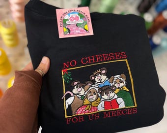 Op de afbeelding: Zwart sweatshirt met een geborduurd ontwerp met cartoonmuizen en de tekst "NO CHEESES FOR US MEEECES" in het rood. Een roze label met tekst is aan het sweatshirt bevestigd.