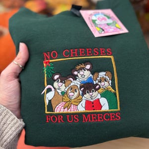 May include: Dark green sweatshirt with embroidered graphic of five mice in a gold frame. The text "NO CHEESES FOR US MEEECES" is embroidered in red. A pink tag is attached to the sweatshirt.