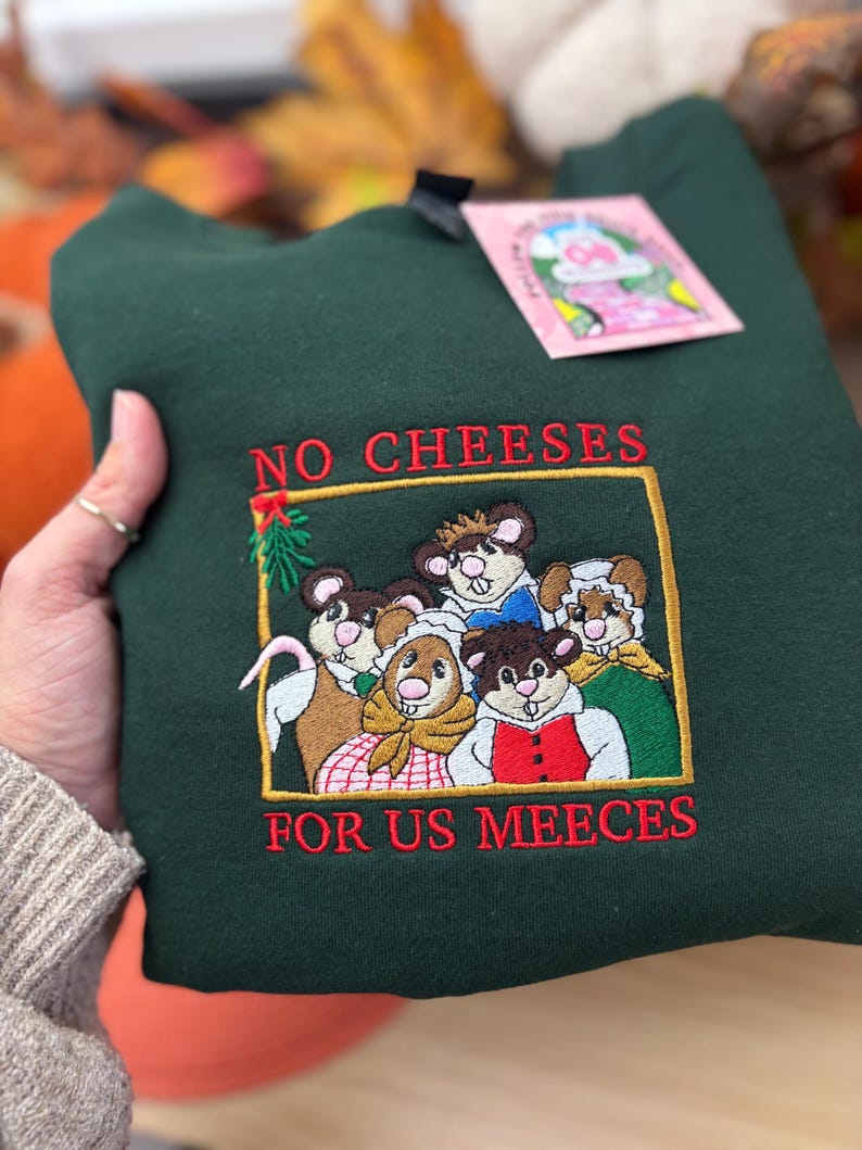 May include: Dark green sweatshirt with embroidered graphic of five mice in a gold frame. The text "NO CHEESES FOR US MEEECES" is embroidered in red. A pink tag is attached to the sweatshirt.