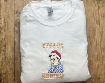 Op de afbeelding: Witte longsleeve t-shirt met een geborduurde afbeelding van een persoon die een kerstmanmuts draagt en de tekst "What a sad little Christmas Jumper, Jane".