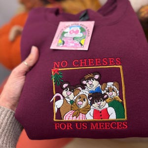 May include: A maroon sweatshirt with embroidered text and a graphic. The text reads "NO CHEESES FOR US MEEECES" in red thread. The graphic features cartoon mice in various outfits. A pink tag is attached to the sweatshirt.