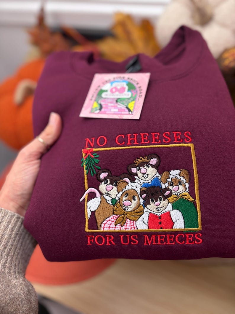 May include: A maroon sweatshirt with embroidered text and a graphic. The text reads "NO CHEESES FOR US MEEECES" in red thread. The graphic features cartoon mice in various outfits. A pink tag is attached to the sweatshirt.