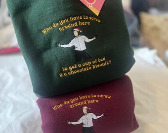 Op de afbeelding: Twee sweatshirts, een groene en een bordeauxrode, met geborduurde tekst die luidt: "Wie moet je lastigvallen om een kopje thee en een chocoladekoekje te krijgen?" De tekst is geel en de afbeelding van een persoon met een kerstmanmuts is wit.