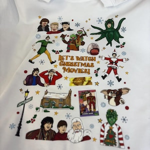 May include: White t-shirt with a festive design featuring characters from Christmas films. The shirt includes the text "Let's Watch Christmas Movies!" and various festive elements like snowflakes and holly.