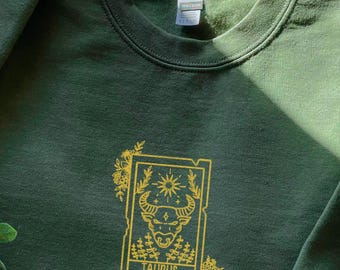 Puede incluir: Una sudadera verde oscuro con un diseño bordado en dorado de una cabeza de toro dentro de un marco con la palabra "TAURUS" debajo. El diseño está rodeado de detalles florales.