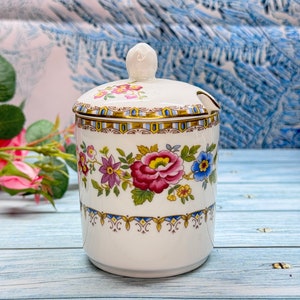 May include: A white ceramic jar with a floral design and a lid. The jar has a gold trim and a blue and white pattern. The lid has a small spoon attached to it.