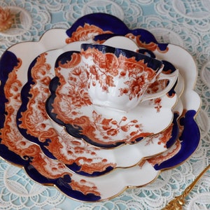 Può includere: Un set di tre tazze da tè e piattini in porcellana bianca con un motivo floreale blu e arancione. I piattini hanno bordi a festoni e le tazze hanno un bordo dorato.