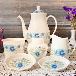 Op de afbeelding: Een set witte keramische keukenartikelen met een blauw bloemenpatroon. De set bevat een theepot, een roomkan, een suikerpot, een pot en twee kleine borden.