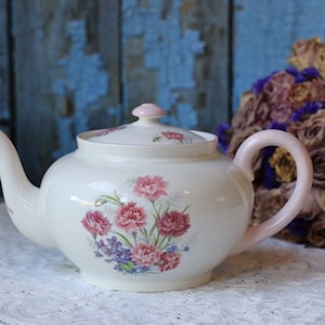 Könnte beinhalten: Eine weiße Teekanne mit einem rosafarbenen Blumendesign und einem rosafarbenen Henkel. Die Teekanne steht auf einer weißen Spitzentischdecke.