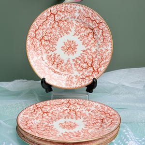 May include: A set of white ceramic plates featuring a detailed orange coral branch pattern across the surface and a thin gold rim. One plate is displayed upright on a black stand, while two identical plates are stacked horizontally below it.