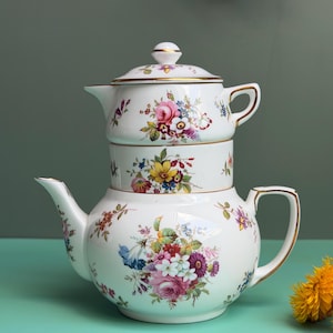 Op de afbeelding: Een wit porseleinen theeservies voor één persoon, bestaande uit een theepot en een gestapeld theekopje met bijpassend deksel. De set is versierd met kleurrijke bloemenboeketten in roze, geel, blauw en groen, afgewerkt met gouden randen.