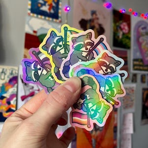 May include: A set of six iridescent holographic stickers featuring a cartoon raccoon holding a pride flag. The flags are in different colours, including rainbow, bisexual, and transgender.