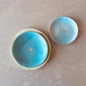 Può includere: Tre piatti in ceramica nei toni del blu e del verde. Il piatto più grande è un turchese vibrante con un motivo a stella dorata. Il piatto centrale è azzurro chiaro e il più piccolo è azzurro pallido con un cuore dorato.