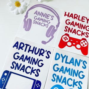 May include: Four white square stickers with gaming-themed designs and text. The stickers feature a purple headset, a red game controller, a blue handheld console, and a blue game controller. Each sticker has a name and the words "Gaming Snacks".