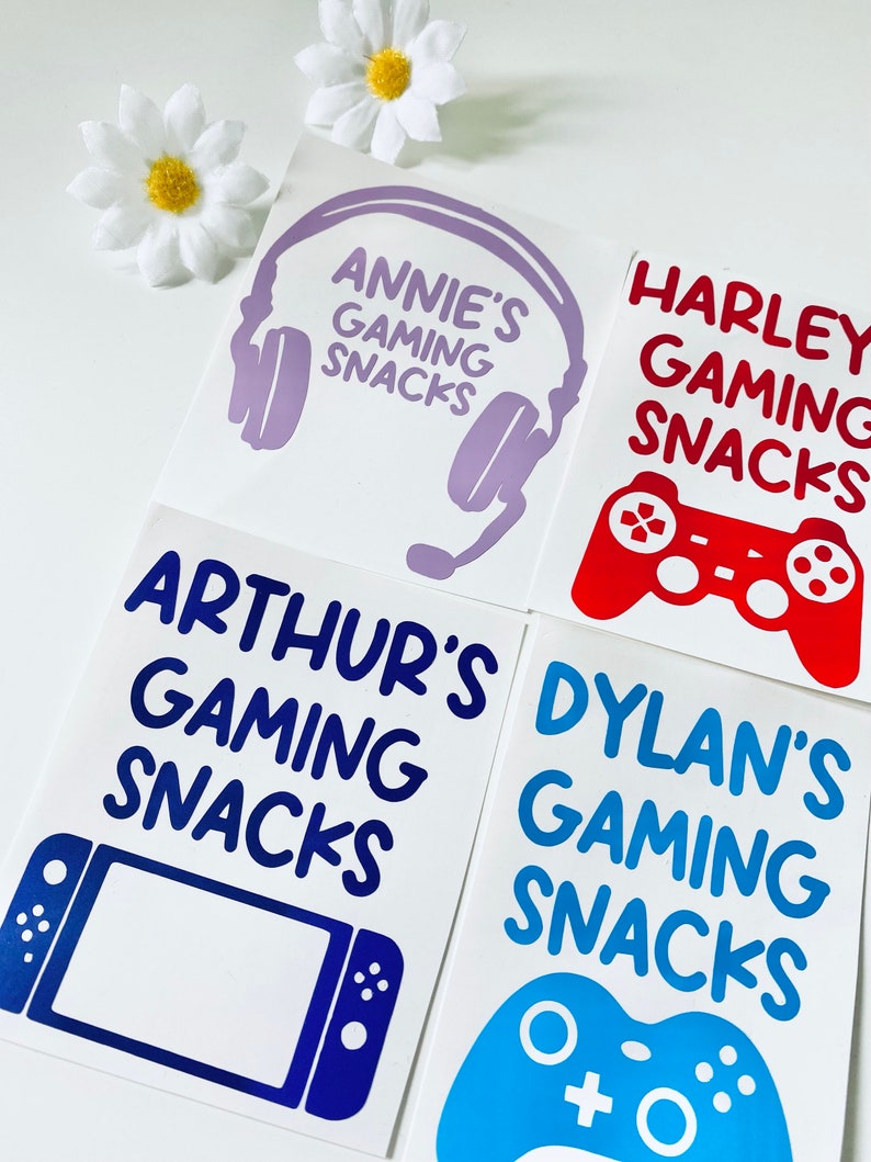 May include: Four white square stickers with gaming-themed designs and text. The stickers feature a purple headset, a red game controller, a blue handheld console, and a blue game controller. Each sticker has a name and the words "Gaming Snacks".