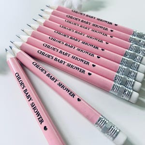 May include: Pink pencils with the text "CHLOE'S BABY SHOWER" printed in black, each with a small black heart. The pencils have silver metal bands and white erasers. A white daisy with a yellow center is in the background.