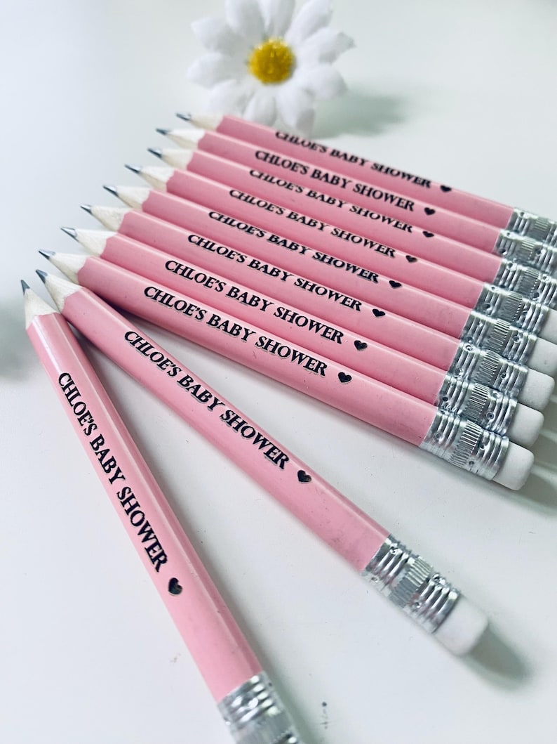 May include: Pink pencils with the text "CHLOE'S BABY SHOWER" printed in black, each with a small black heart. The pencils have silver metal bands and white erasers. A white daisy with a yellow center is in the background.