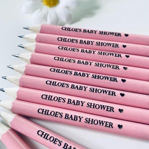 May include: Pink pencils with the text "CHLOE'S BABY SHOWER" printed in black. Each pencil has a small black heart symbol. A white daisy with a yellow center is in the background. The pencils are sharpened and ready for use.