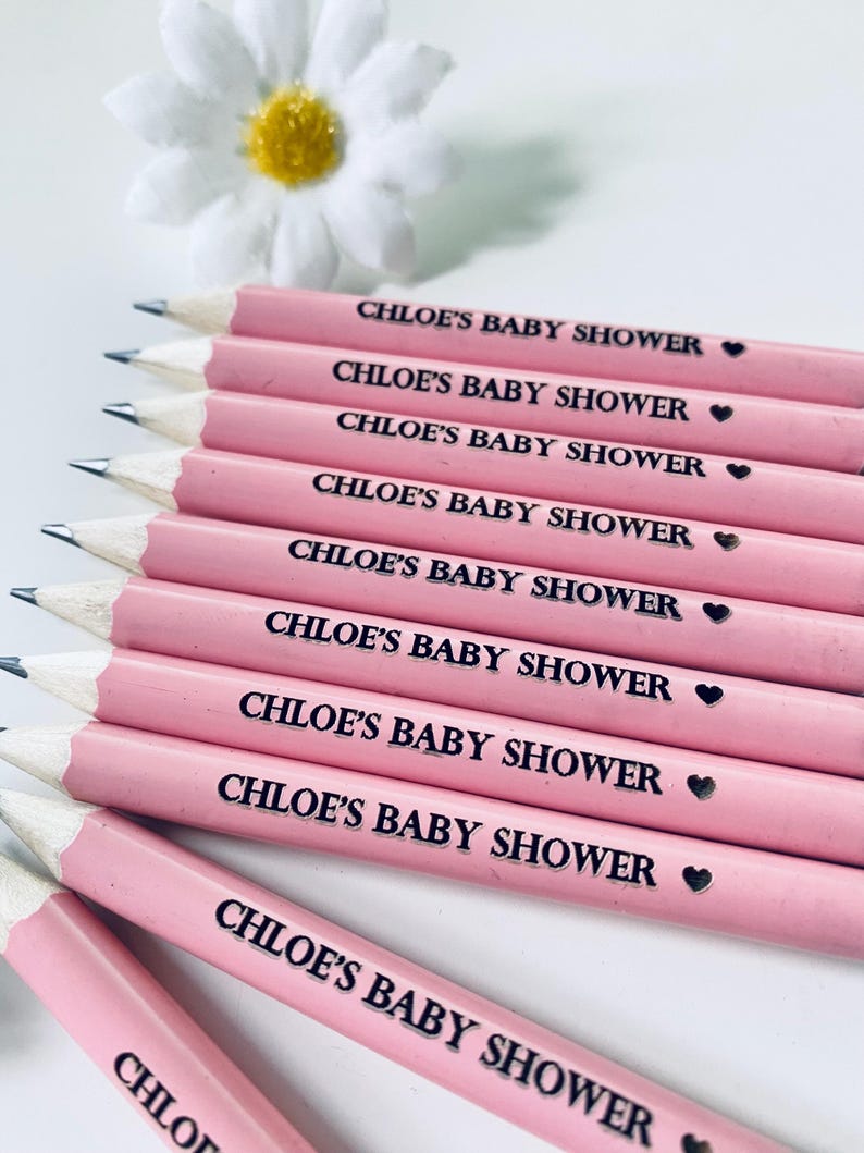 May include: Pink pencils with the text "CHLOE'S BABY SHOWER" printed in black. Each pencil has a small black heart symbol. A white daisy with a yellow center is in the background. The pencils are sharpened and ready for use.