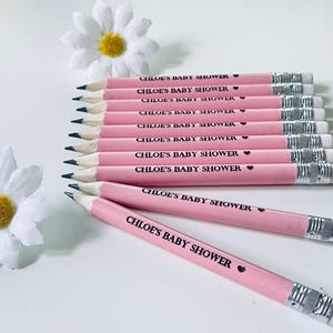 May include: A collection of pink pencils with the text "CHLOE'S BABY SHOWER" printed in black. Each pencil has a silver ferrule and a white eraser. Two white daisy flowers are in the background.