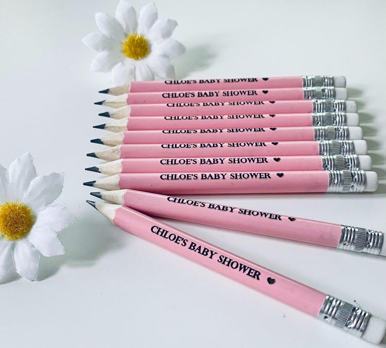 May include: A collection of pink pencils with the text "CHLOE'S BABY SHOWER" printed in black. Each pencil has a silver ferrule and a white eraser. Two white daisy flowers are in the background.