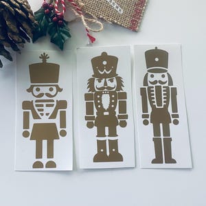 May include: Three gold-coloured nutcracker stickers on white backing. Each sticker features a different design of a traditional nutcracker soldier. The stickers are arranged in a row, with a festive holiday theme.