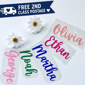 May include: Five clear adhesive vinyl decals featuring the names Olivia in coral, Ethan in magenta, Martha in blue, Noah in green, and George in light pink, all written in a cursive script font, accompanied by two white artificial daisy flowers.