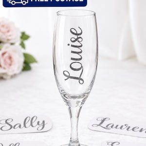 May include: A clear glass champagne flute featuring the name Louise in a dark grey cursive script. The glass is surrounded by several white oval labels, each printed with a different name in the same dark grey cursive font style.
