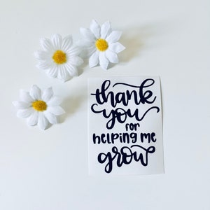 May include: A white card with black calligraphy that reads "thank you for helping me grow". Three white daisies with yellow centres are arranged around the card.