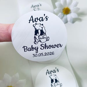 May include: White, circular stickers featuring a Winnie the Pooh design and the text "Ava's Baby Shower 30.05.2026". The stickers are on a roll, with a daisy in the background.
