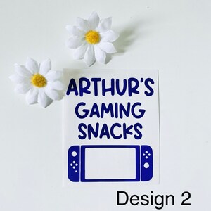 May include: A white sticker with the text "ARTHUR'S GAMING SNACKS" in blue, along with a blue game console graphic. Two white daisy flowers with yellow centers are also present. The design is labeled "Design 2".