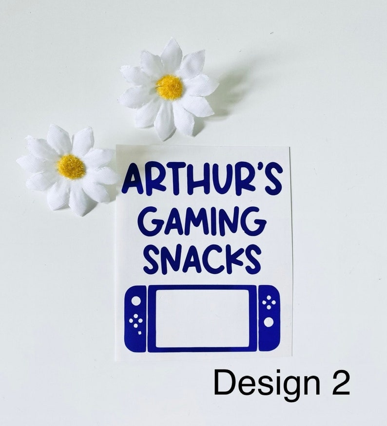 May include: A white sticker with the text "ARTHUR'S GAMING SNACKS" in blue, along with a blue game console graphic. Two white daisy flowers with yellow centers are also present. The design is labeled "Design 2".