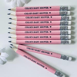 May include: A collection of pink pencils with the text "CHLOE'S BABY SHOWER" printed in black, each accompanied by a small heart symbol. The pencils have silver metal bands and white erasers. The pencils are arranged on a white surface.