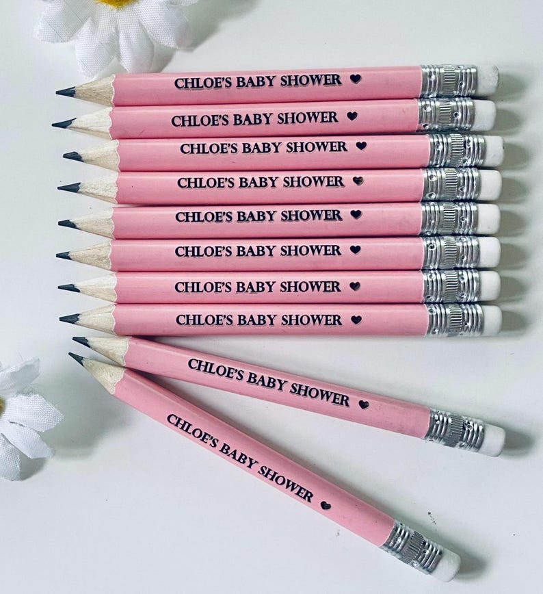 May include: A collection of pink pencils with the text "CHLOE'S BABY SHOWER" printed in black, each accompanied by a small heart symbol. The pencils have silver metal bands and white erasers. The pencils are arranged on a white surface.