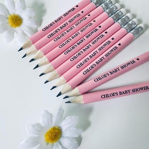May include: Pink pencils with silver ferrules and black tips, arranged on a white surface. Each pencil is printed with the text "CHLOE'S BABY SHOWER" and a small heart. White daisy flowers with yellow centers are in the background.