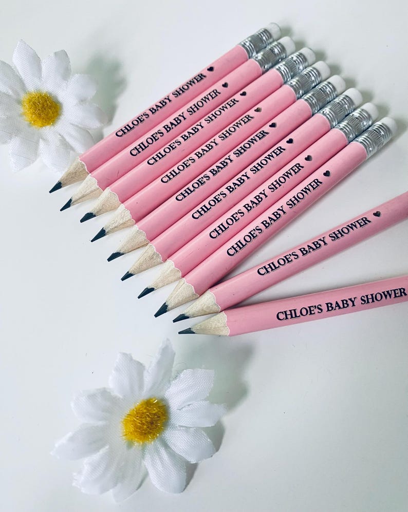 May include: Pink pencils with silver ferrules and black tips, arranged on a white surface. Each pencil is printed with the text "CHLOE'S BABY SHOWER" and a small heart. White daisy flowers with yellow centers are in the background.