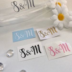 May include: Five different versions of a monogram sticker with the letters "S&M" in a cursive font. The stickers are on a white background. The stickers are in black, white, gold, light blue, and pink.