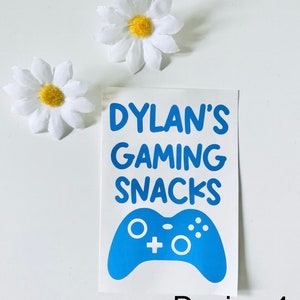 May include: A white sticker with blue text that reads "Dylan's Gaming Snacks" and a blue game controller graphic. Two white daisy flowers with yellow centers are also present. The sticker design is labeled "Design 4".