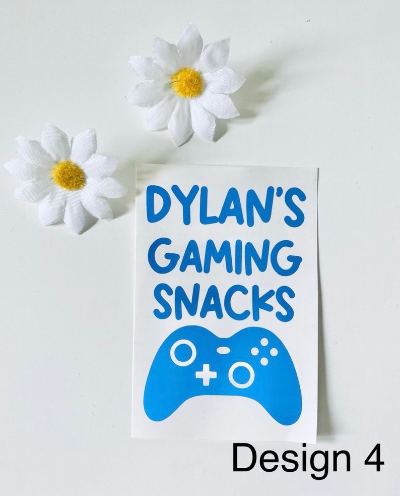 May include: A white sticker with blue text that reads "Dylan's Gaming Snacks" and a blue game controller graphic. Two white daisy flowers with yellow centers are also present. The sticker design is labeled "Design 4".