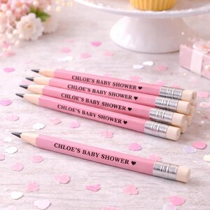 May include: Pink pencils with the text "CHLOE'S BABY SHOWER" are arranged on a light wood surface, with cupcakes on a white cake stand in the background. Pink confetti and a small gift box are also present.