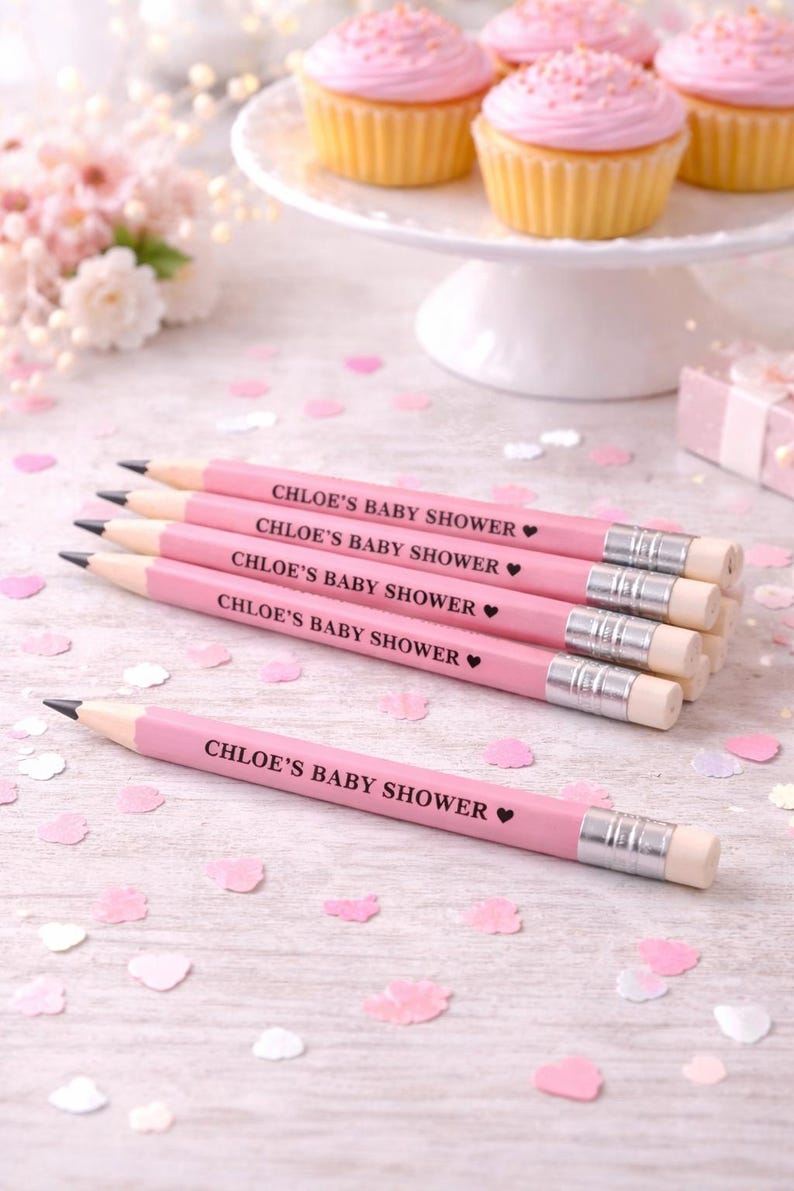 May include: Pink pencils with the text "CHLOE'S BABY SHOWER" are arranged on a light wood surface, with cupcakes on a white cake stand in the background. Pink confetti and a small gift box are also present.
