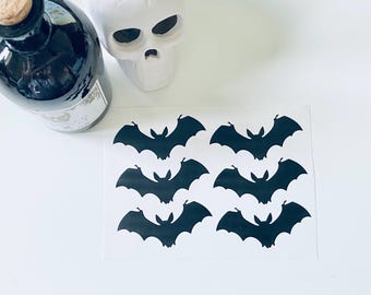Puede incluir: Naturaleza muerta con temática de Halloween con una botella negra con corcho, una calavera blanca y una hoja de pegatinas de murciélagos negros. Los murciélagos son simétricos.