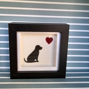 May include: A framed picture with a black silhouette of a dog and a red heart. The artwork is set against a white background within a black frame. The background is a blue and white striped pattern.